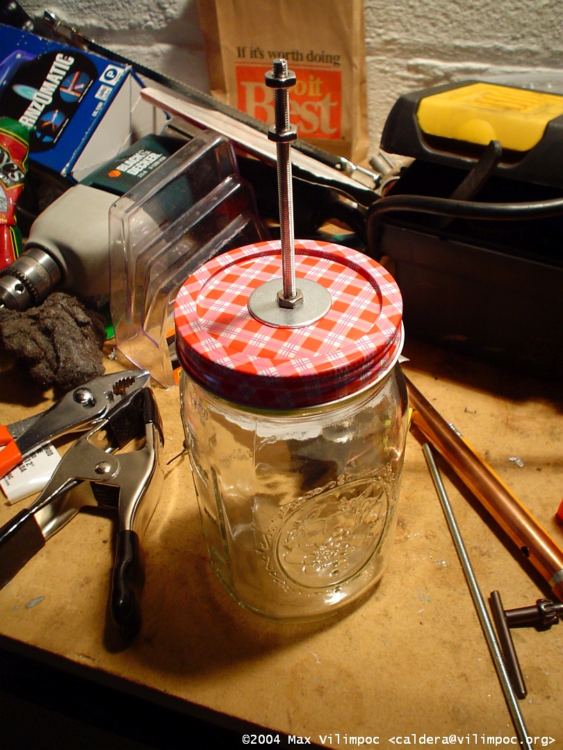 A glass mason jar, empty, with a 6 inch piece of one quarter inch rebar bolted into the lid with hex nuts and one inch washers. Next to the jar on the workbench is the chuck from a drill, a Black and Decker corded drill, a pair of pliers, and a spring-loaded clamp