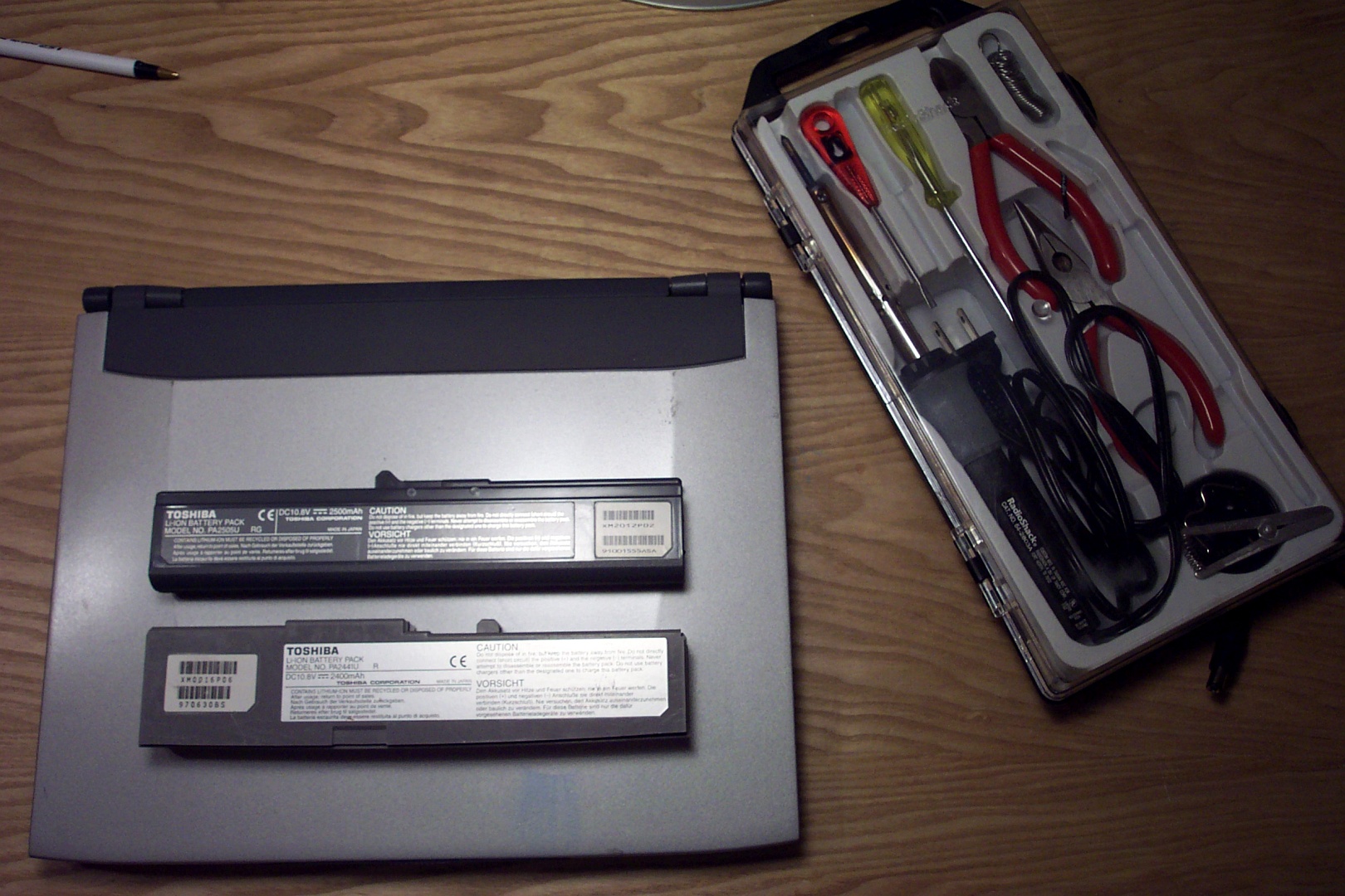 Two laptop batteries, unopened, sitting next to an electronics toolkit containing a soldering iron, screwdrivers, wirecutters, and a spool of solder