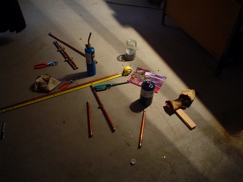 A view of a bunch of tools on the floor of the basement workspace, pliers, copper tubing, the propane torch, a can of propane, a barbecue lighter, and leather gloves