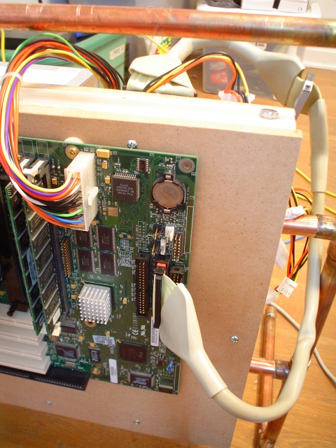 A closeup of the microATX motherboard mounted to the middle-density fiberboard, with the whole machine standing, and ATX power cable and parallel IDE cable visible